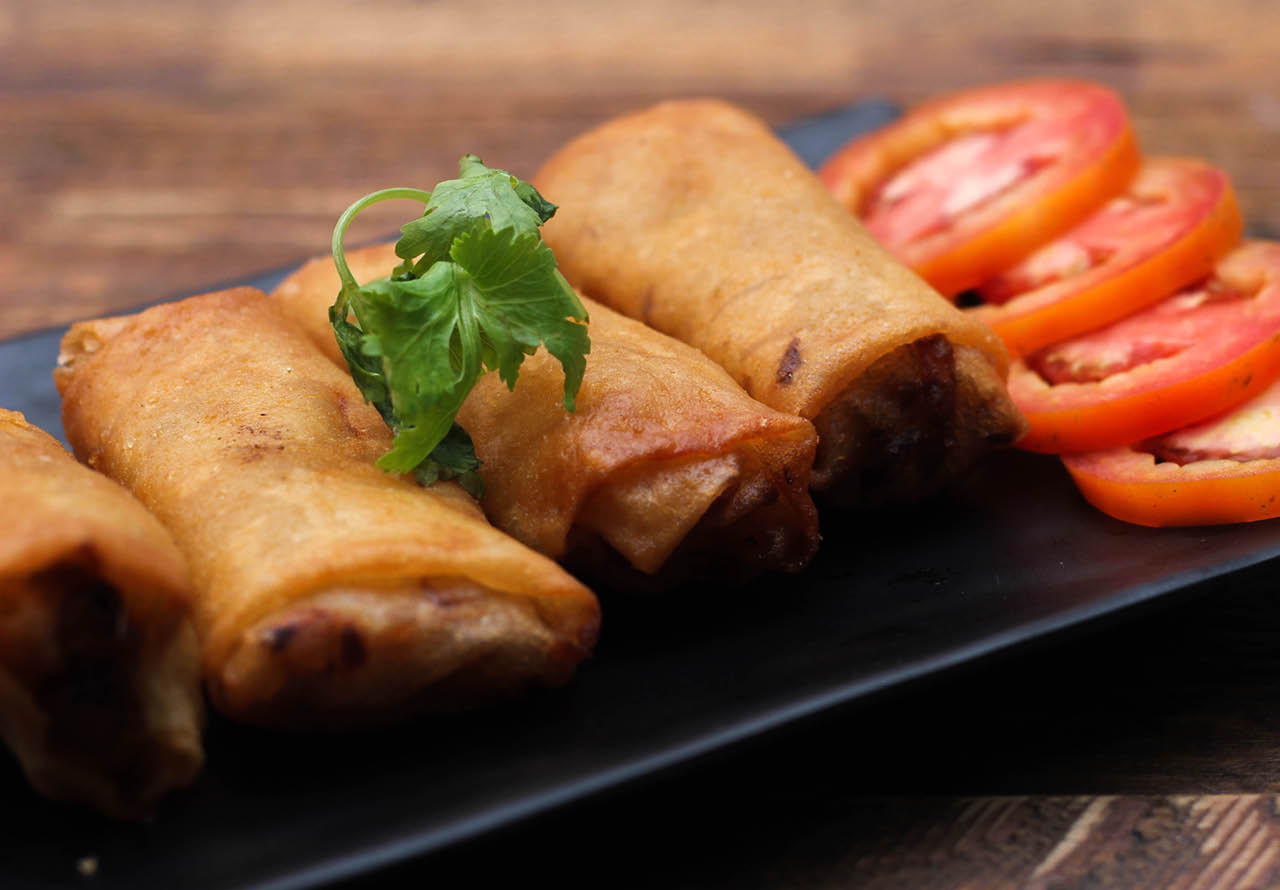 Fried spring rolls served in a black square plate with sliced tomatoes on a brown wooden table in Thailand.